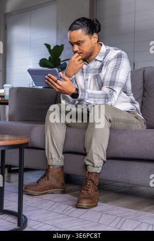 Adult man sitting on gray sofa holding tablet in folio and touching chin in living room Stock Photo