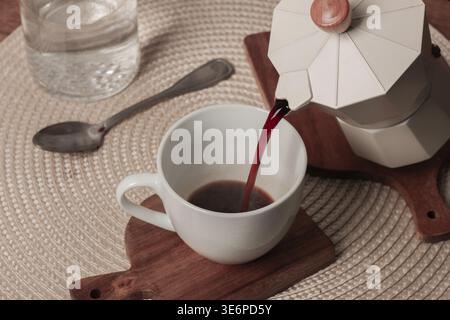 A coffee maker pours steaming coffee into a white cup placed on a wooden mat. Nearby, a glass of water and a spoon complete the cozy breakfast setting Stock Photo