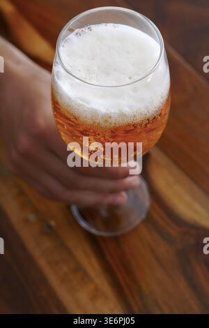 Hand holding a cold glass of light golden beer with frothy white head and lively bubbles on a natural wooden tabletop, top view capturing refreshing, Stock Photo