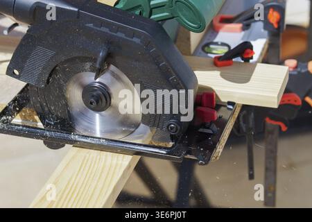 Circular saw blade actively cutting through a wooden plank, creating sawdust and demonstrating carpentry work on a workbench with other tools and clam Stock Photo