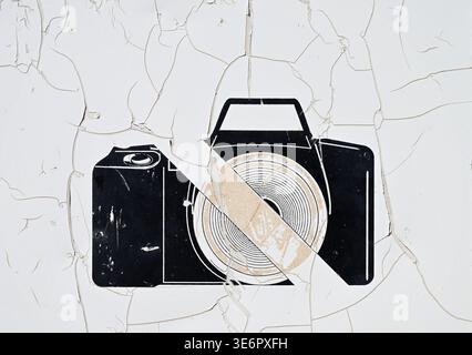 The image shows a stylized black camera icon painted on a white wall whose surface is heavily cracked and peeling. A diagonal stripe runs across the c Stock Photo
