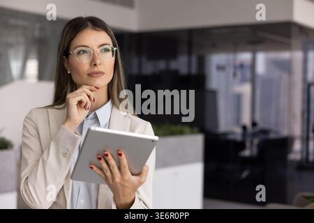 Thoughtful professional woman stands in modern office with tablet Stock Photo