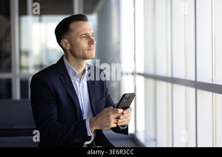 Serious European businessman holds smartphone, look out office window thoughtfully, make mobile phone call to client, thinking on response at workplac Stock Photo