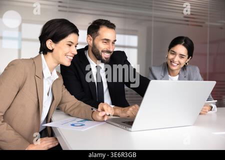 Smiling business team gathered around laptop, collaborating on project, reviewing financial data, working on strategy together in office. Effective te Stock Photo