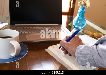 Person's hand writing in a planner with laptop and coffee, symbolizing home office productivity Stock Photo