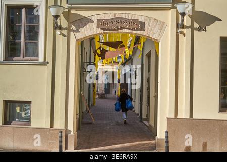 A sunlit stone archway entrance to a narrow pedestrian alleyway labeled "skersvėja" with decorative yellow streamers and a metal silhouette of a Stock Photo