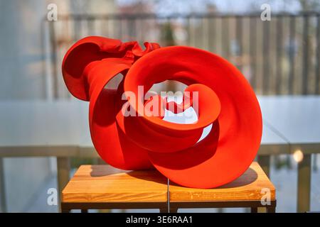 A vibrant red abstract sculptural artwork displayed on a wooden pedestal stand. The sculpture features smooth, flowing organic forms with twisted Stock Photo