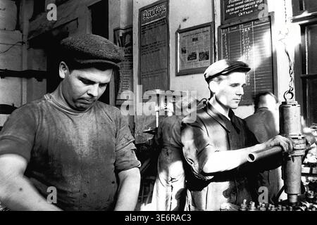 Sloviansk, 1960s. Inside the reinforcement shop of the Armature and Insulator Plant (AIZ). A Soviet worker in a cap operates a heavy drilling machine at his workplace, while his colleague stands nearby focused on a task. The industrial interior features a bulletin board and safety instructions on the wall. This authentic black and white archival photo captures daily manual labor, factory manufacturing processes, and the industrial atmosphere of a bygone era, reflecting the history of peaceful Donbas. Stock Photo