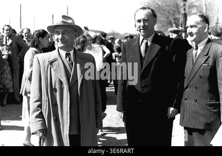 Sloviansk, 1950. Outside the Armature and Insulator Plant (AIZ). Three distinguished Soviet men, likely plant management or senior factory engineers, stand confidently together in formal suits and coats. They are participating in a lively outdoor public gathering just before the start of a festive mass demonstration. A bustling crowd of local citizens is visible in the bright background. This authentic black and white archival photograph captures community leadership and the historical memory of peaceful Donbas. Stock Photo