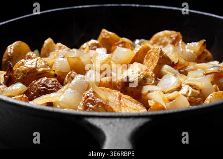 Fried crispy chopped potato and onion cooked in a cast iron frying pan. On a black background backdrop Stock Photo