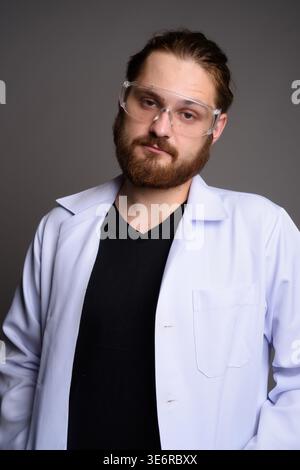 Studio portrait of a bearded young man in a white lab coat and protective safety goggles looking at the camera with a serious expression. Concept for Stock Photo