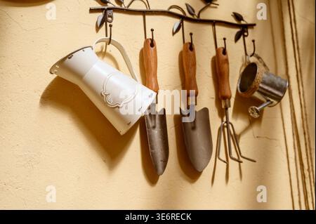 White decorative pitcher and mini rake hanging in a cozy garden setting on a wall. Concept of domestic plant cultivation tools and aesthetic yard main Stock Photo