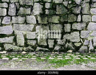 A high-quality texture shot of a rustic stone wall built from irregular granite blocks with dark crevices and patches of green moss Stock Photo