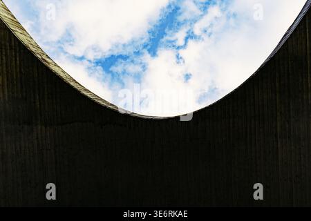 A striking architectural composition featuring a dark, textured concrete underpass arch that creates a dramatic silhouette against a bright blue sky Stock Photo