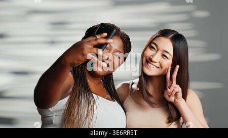 Diverse friends taking photos on mobile phone in studio, expressing self love and self confidence. Young beauty models promoting body positivity and different skintones, wellness. Stock Photo