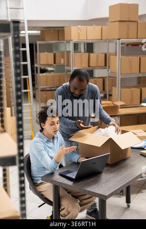 Diverse workers checking digital inventory system for delivery logistics, using laptop and coordinating shipment services in a warehouse setup. Local brand operations and e commerce growth. Stock Photo