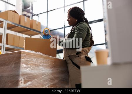 African american supevisor taping clients orders using adhesive tape in storehouse, preparing packages for shipping. Stockroom employee working at merchandise quality control in warehouse Stock Photo