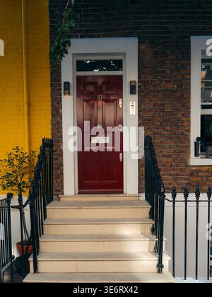 London, United Kingdom - 21 June 2025: View of a vibrant red door framed by stark white trim, steps leading up, the dark railings contrasting against Stock Photo