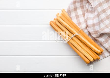 A bundle of golden grissini breadsticks tied with twine rests on a beige checkered napkin atop a white wooden table, evoking rustic snack, Italian bak Stock Photo