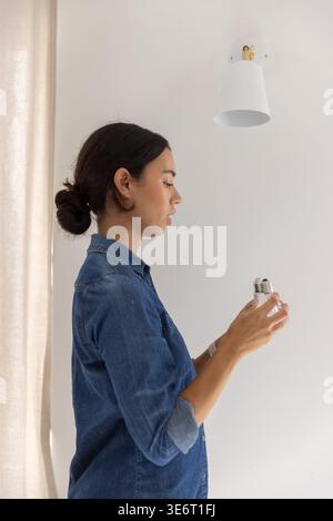 LED lightbulb preparing to be installed into empty lamp socket on white wall near beige curtain Stock Photo