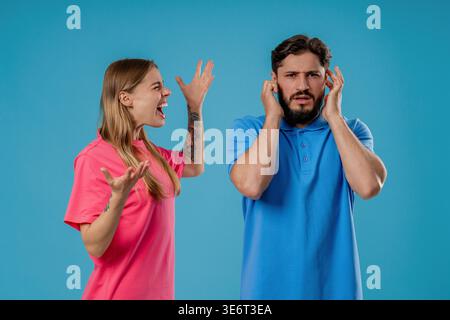 Young woman screaming on husband or boyfriend On blue. Tired man shut ears. Concept of conflict, quarrel, problems in couple relationships. High quality Stock Photo