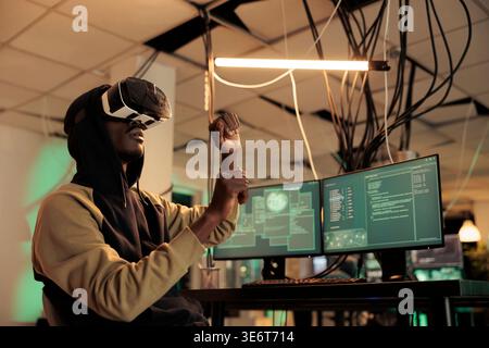 African american hacker working at night with 3d vr glasses, trying to break in government server with virtual reality. Male criminal using interactive headset to steal sensitive official data. Stock Photo