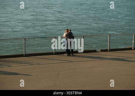 Rear view of a couple embracing while standing at a metal railing, looking out over a wide river during the warm light of a late afternoon. Stock Photo