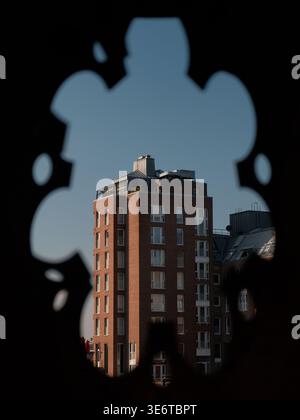 Red brick apartment building seen through a dark, ornate silhouette frame, featuring a rooftop terrace and many windows against a clear blue sky. Stock Photo