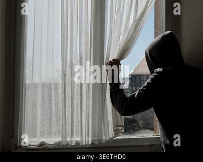 A figure in a hoody pushes the curtain aside, looking out of a window. Stock Photo