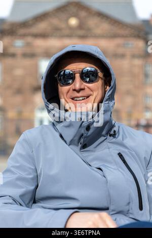 Smiling traveler wearing hooded jacket sunglasses enjoy sunny day European city historic architecture background. Reflections in glasses show lively Stock Photo