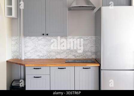 Empty modern minimal tiny grey color kitchen, marble wall, solid natural wood butcher block countertop with built in small stove, black handles. Stock Photo