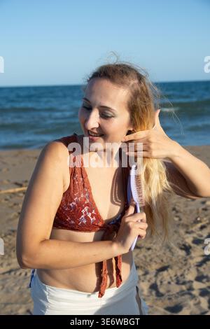Young woman smiling and brushing her long blonde hair on a sandy beach by the ocean, enjoying a moment of self care and relaxation during her summer v Stock Photo