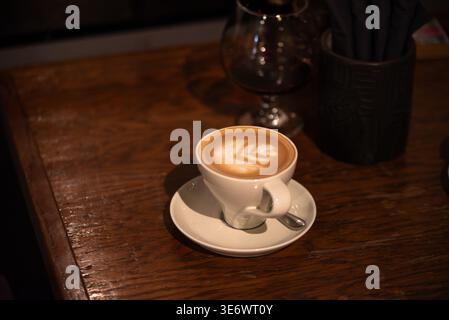 A cup of cappuccino with latte art sits on a white saucer with a spoon, on a wooden table in a cozy, dimly lit café. Stock Photo