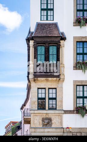 Ornate bay window on historic white building with carved stonework, dark wooden upper window, green-framed panes and flower boxes, clear blue sky Stock Photo