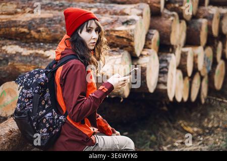 Woman with backpack sits on log while holding smartphone in front of stacked timber. hiking navigation app, mountain gps tracking, digital map Stock Photo