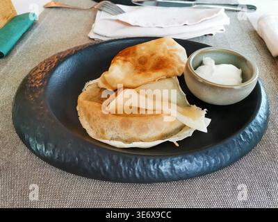 Two pieces of traditional pancake served on a black plate alongside a small bowl of yogurt dip, arranged on a textured table setting with utensils vis Stock Photo