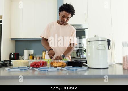 Adult African American man chopping greens at kitchen island, cutting board, blue lids, pink tee Stock Photo