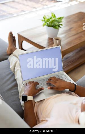Adult African man reclining on gray sofa, typing on silver laptop with white smartwatch visible Stock Photo