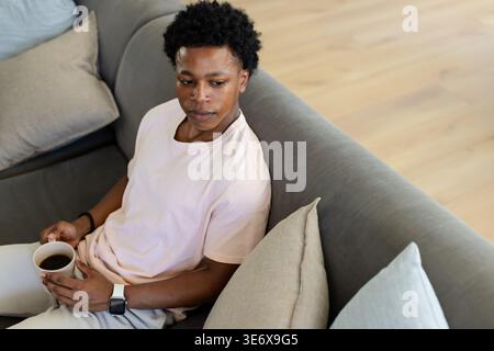 African American man sitting on gray sofa at home holding white coffee mug and wearing smartwatch Stock Photo