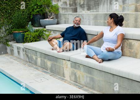 Senior African American couple sitting on padded bench at backyard swimming pool, holding hands Stock Photo