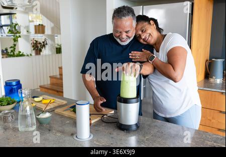 Senior couple smiling and preparing green smoothie at home kitchen island using blender and avocado Stock Photo