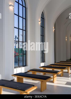 Peaceful interior of a modern minimalist white chapel or ceremonial hall featuring large arched windows, sunlight casting shadows, and rows of simple Stock Photo