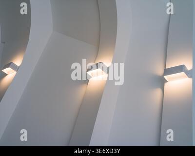 Abstract perspective of smooth white curved architectural walls illuminated by modern square wall sconces glowing with warm light. The scene represent Stock Photo