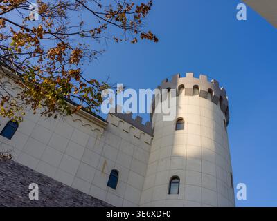 Low angle view of a classic white castle-style round tower with crenellations, partially framed by autumn tree branches under a clear blue sky. The sc Stock Photo