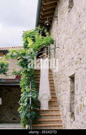 Elegant Dress Hangs By Weathered Wall. Sunlight Creates Warm Contrast On Courtyard Bridal Gown Stock Photo