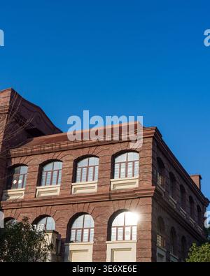 Low angle view of a classic red brick architectural facade featuring large arched glass windows reflecting bright sunlight under a clear blue sky. The Stock Photo