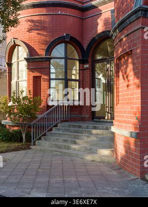 Classic red brick building entrance featuring a stone staircase, arched glass doors, and paved walkways in a sunny outdoor setting. The scene represen Stock Photo