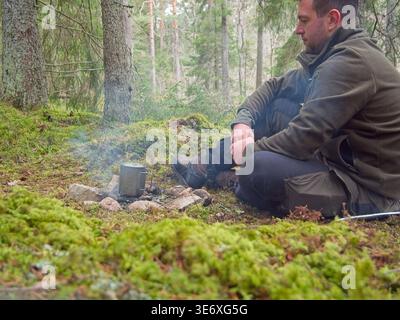 Man resting by campfire during bushcraft camping in forest Stock Photo