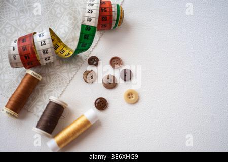 A sewing composition featuring a coiled measuring tape, spools of thread in sha-des of brown and gold, and assorted buttons on a light, textured backg Stock Photo