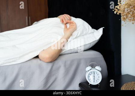 A woman lies in bed with her arm raised under a white blanket, an alarm clock ringing on the bedside table beside her, in a dark room with a wooden wa Stock Photo